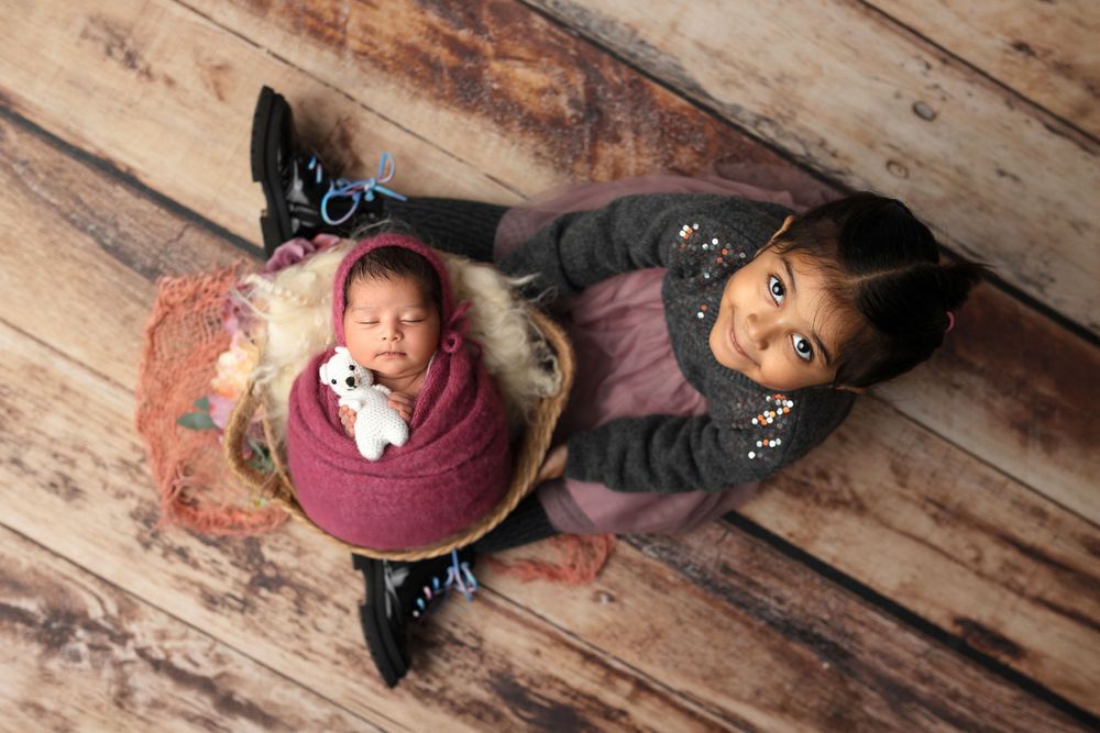 Top view shot of a baby wrapped in burgundy swaddle lying on wooden floor next to sibling.