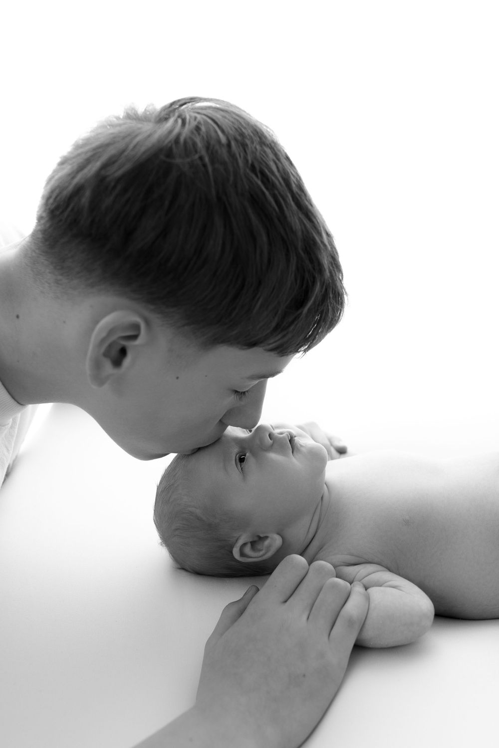 A tender black and white portrait of a child leaning over to kiss a newborn baby lying on white fabric.