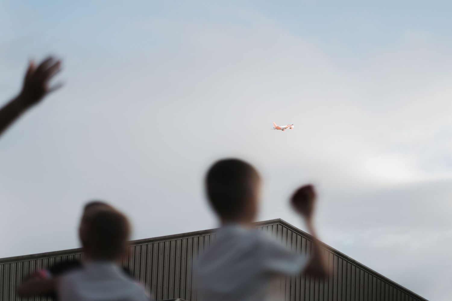 Silhouetted figures watching an airplane in the cloudy sky above a metal building.