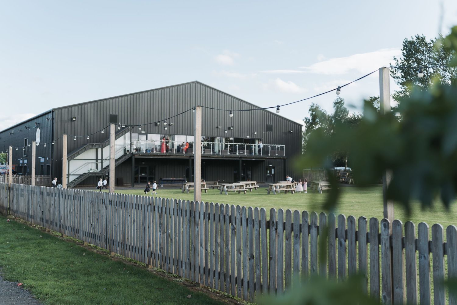 A metal industrial building with a raised viewing deck sits behind a gray picket fence on a grassy field.