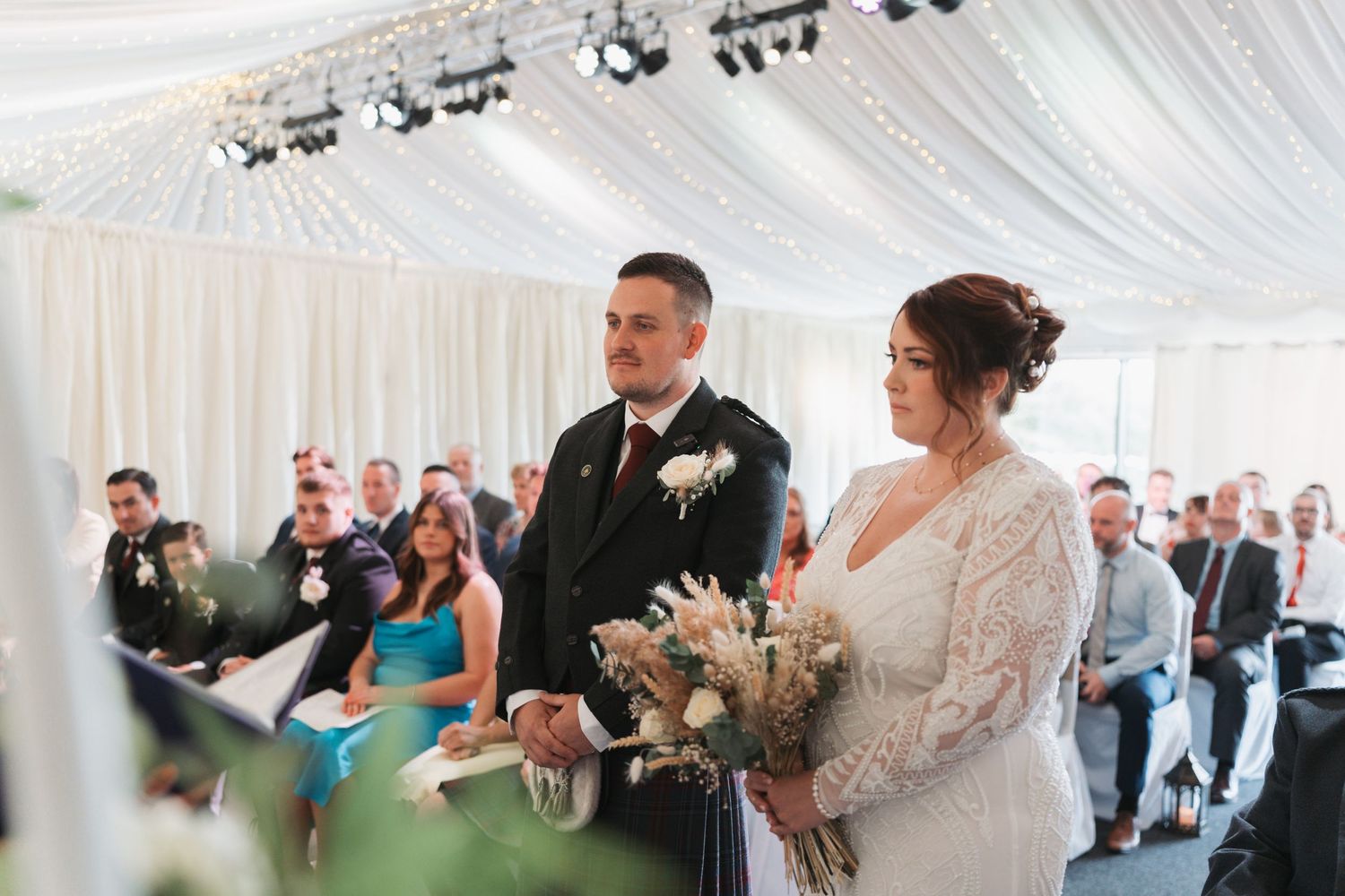 A couple stands together during their indoor wedding ceremony under a white tent with guests seated in the background.