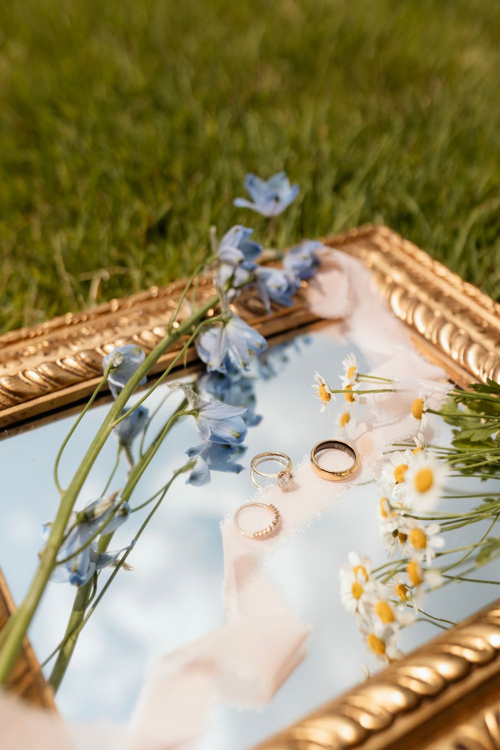 Gold-framed mirror reflects wedding rings, delicate blue flowers and daisies arranged on light surface.
