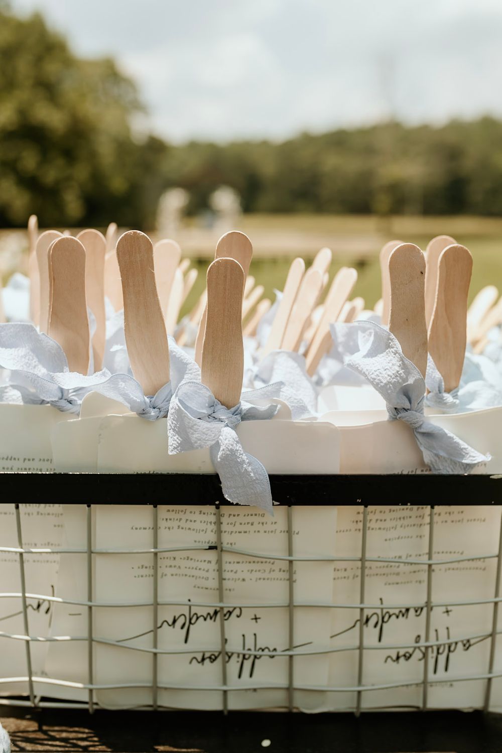 White paper programs with wooden popsicle sticks arranged in a rustic basket for an outdoor wedding event.