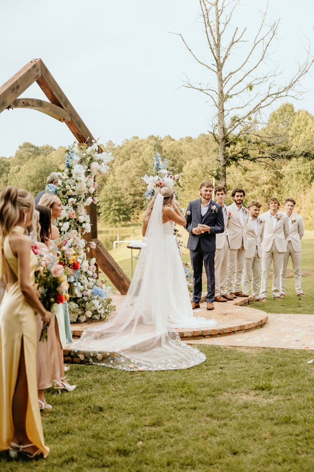 A rustic outdoor wedding ceremony under a wooden arch decorated with flowers and greenery on a grassy lawn.