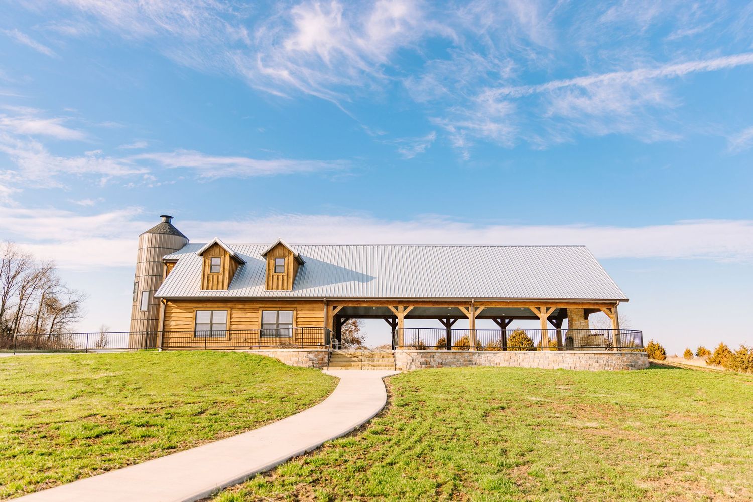 A rustic yellow barn with a covered archway and silo sits atop a rolling green hill under bright blue skies.