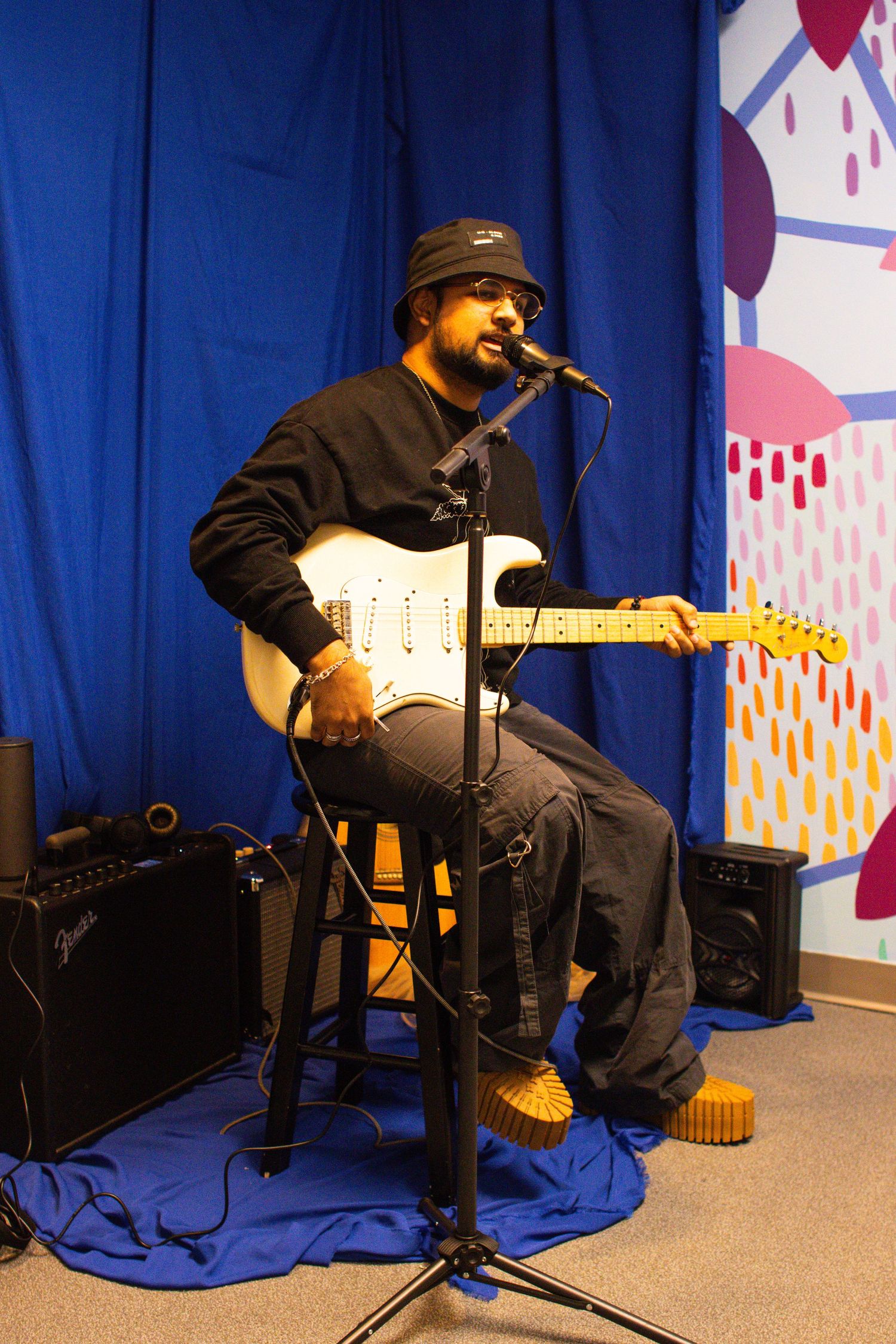 A musician performs on electric guitar while seated against a blue curtain and patterned background.