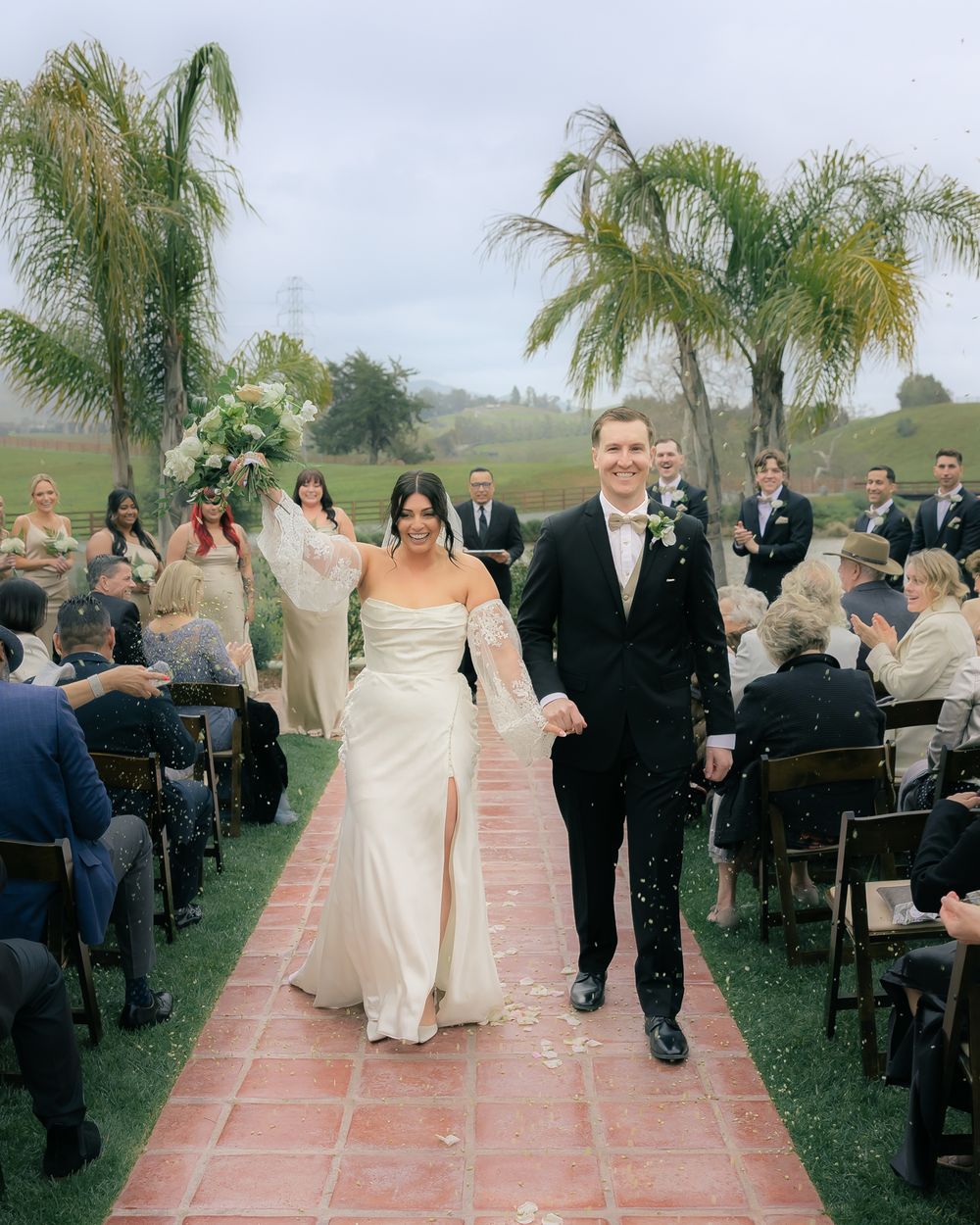 A wedding ceremony processional on a tiled pathway surrounded by palm trees.