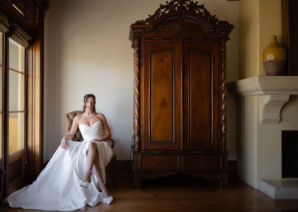 A person in a white wedding dress sits elegantly beside an ornate vintage wooden armoire in a sunlit room.