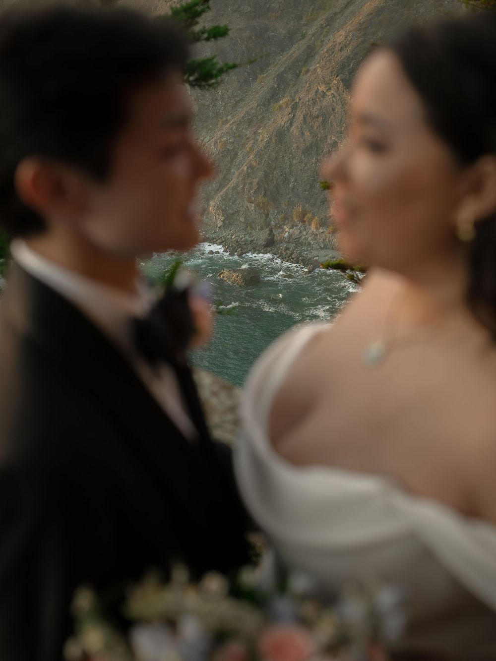 Blurred wedding couple in formal attire with scenic coastal cliffs and turquoise water in the background.