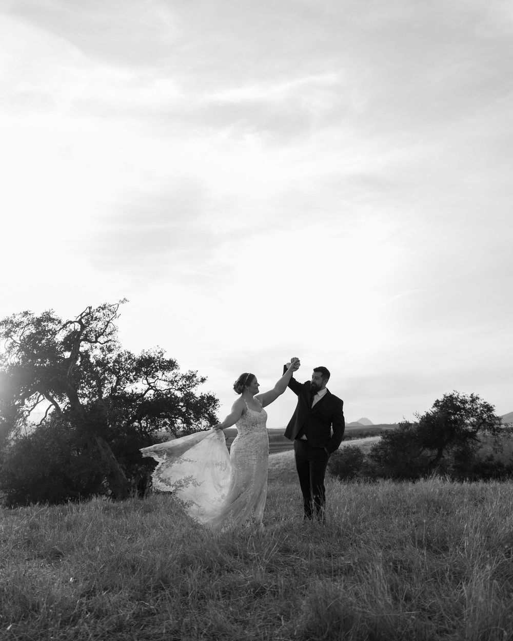 Black and white photo of a bride and groom dancing in a field at sunset, her gown flowing in the wind.