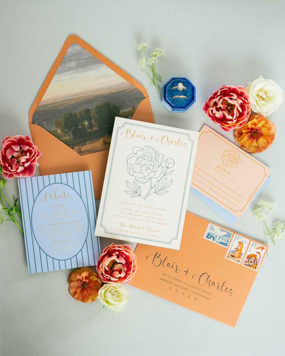 Elegant wedding invitation suite in peach and blue with floral details, envelope liner art, and fresh blooms on a gray background.