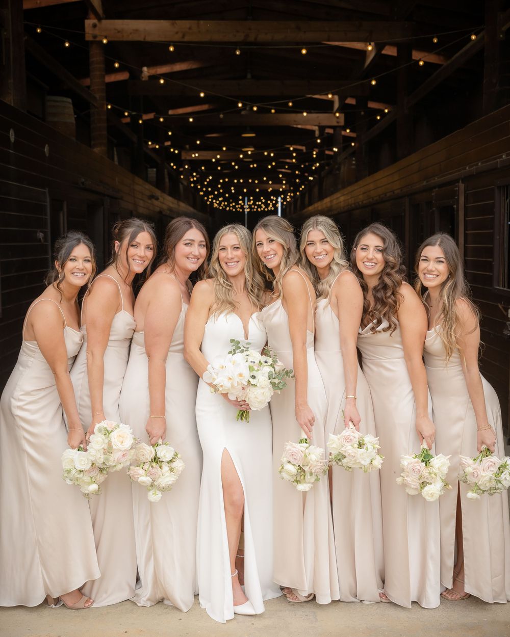 Bridal party wearing champagne-colored silk gowns holding white bouquets pose in a rustic venue with twinkle lights overhead.