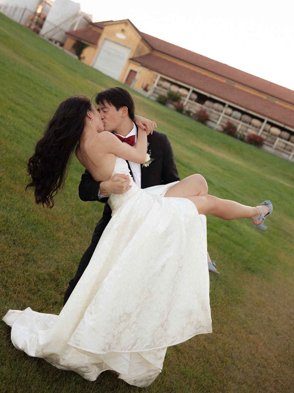 Groom lifting bride in white wedding dress for a romantic kiss outdoors near a barn venue on green grass.