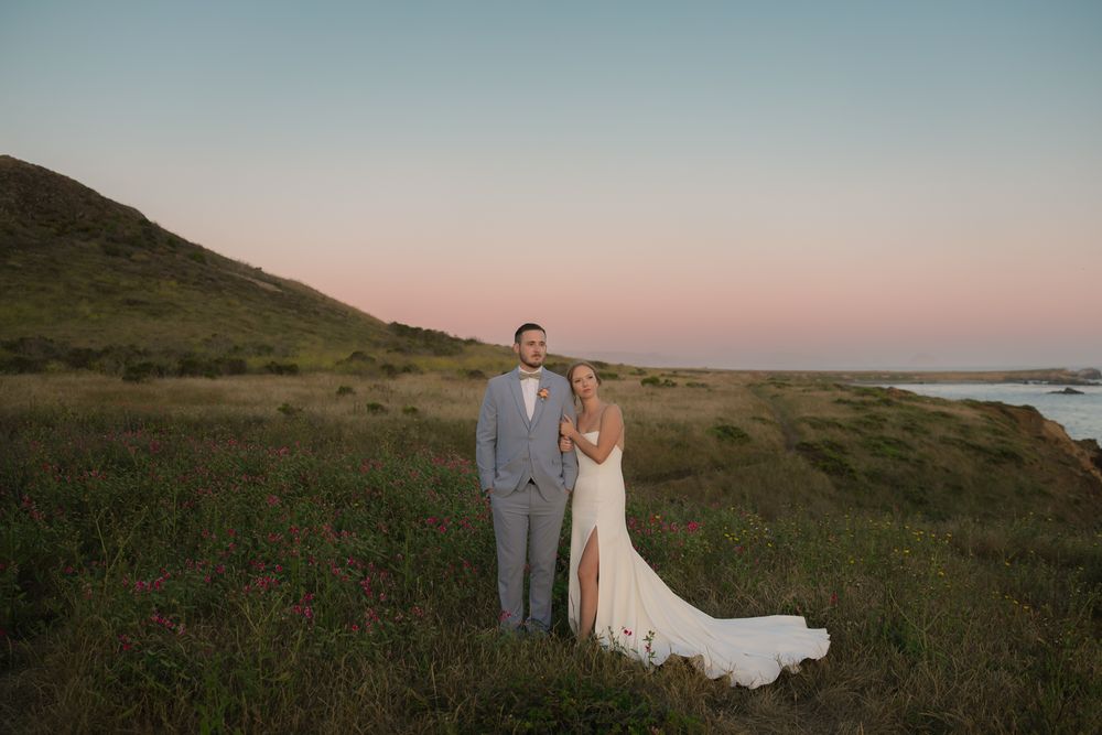 Bride and groom pose in a lush green meadow at sunset with a pink sky and coastal hills backdrop.