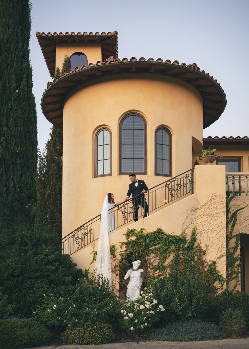 Bride and groom on ornate staircase of Tuscan-style villa with arched windows and lush garden.