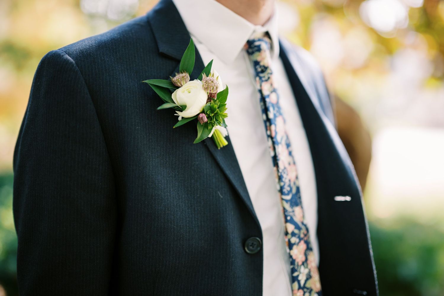 A white rose and greenery boutonniere pinned to a navy blue suit jacket with a floral patterned tie.