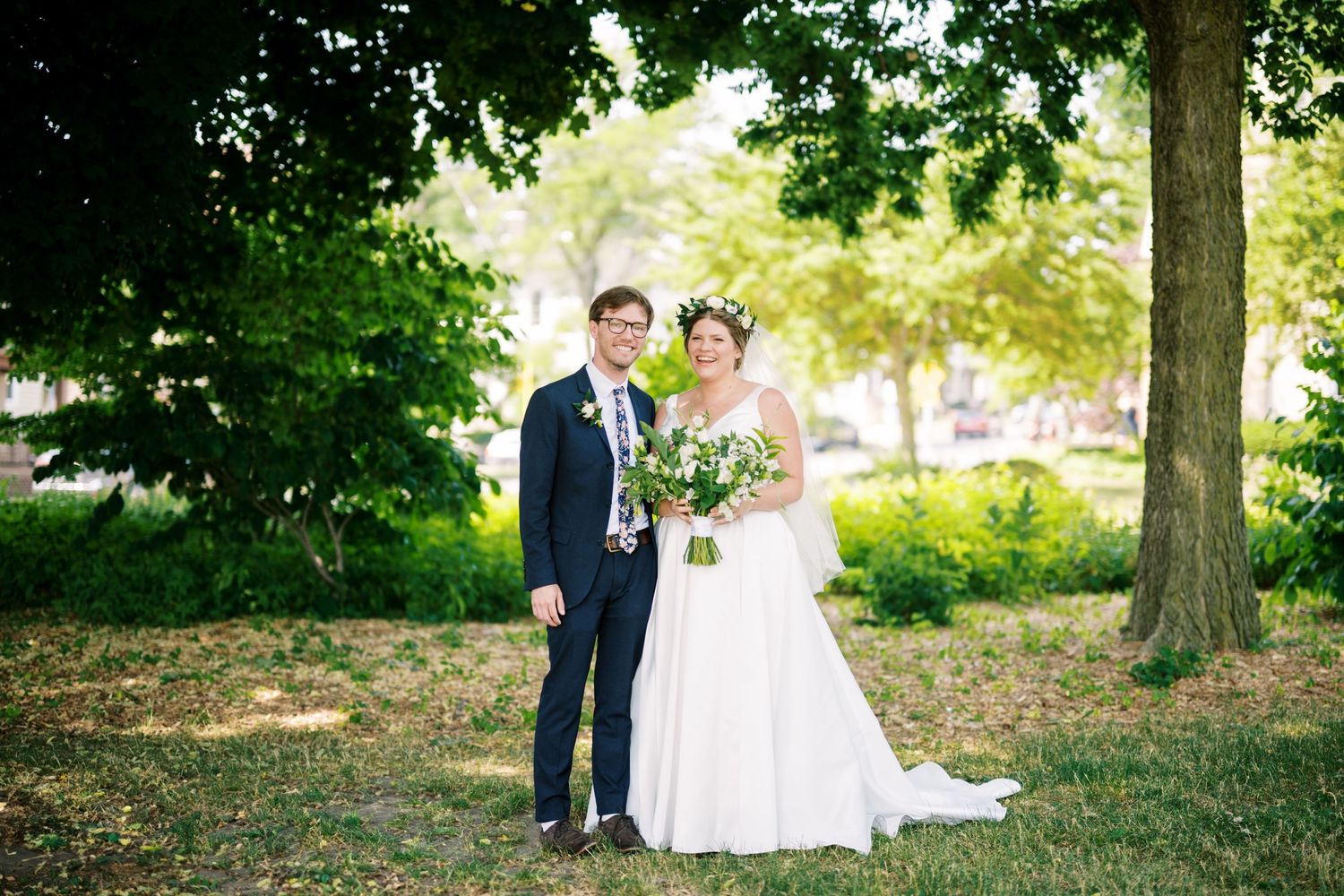 A couple poses for a wedding photo outdoors in a park setting with green trees and natural lighting.