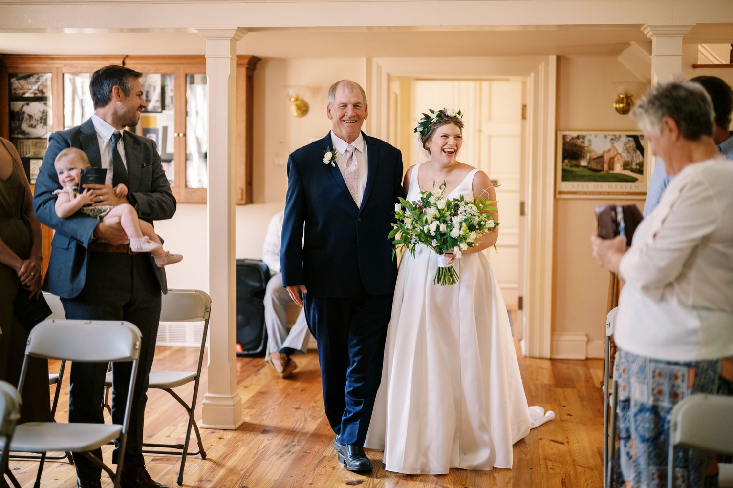 A wedding processional takes place in a rustic indoor venue as people photograph the special moment.