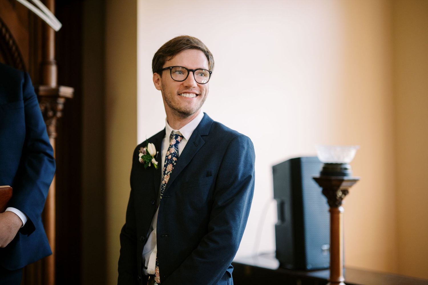 A smiling person in a navy suit and glasses stands beside a wall during a formal event.
