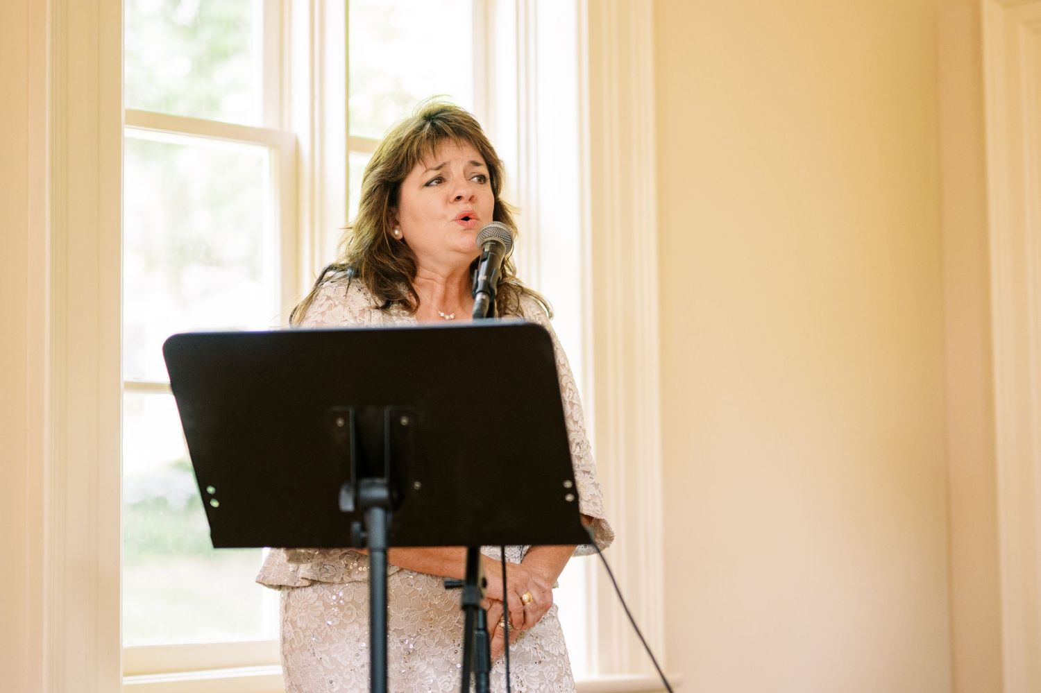 A vocalist performs with a music stand by a sunlit window in a room with beige walls.