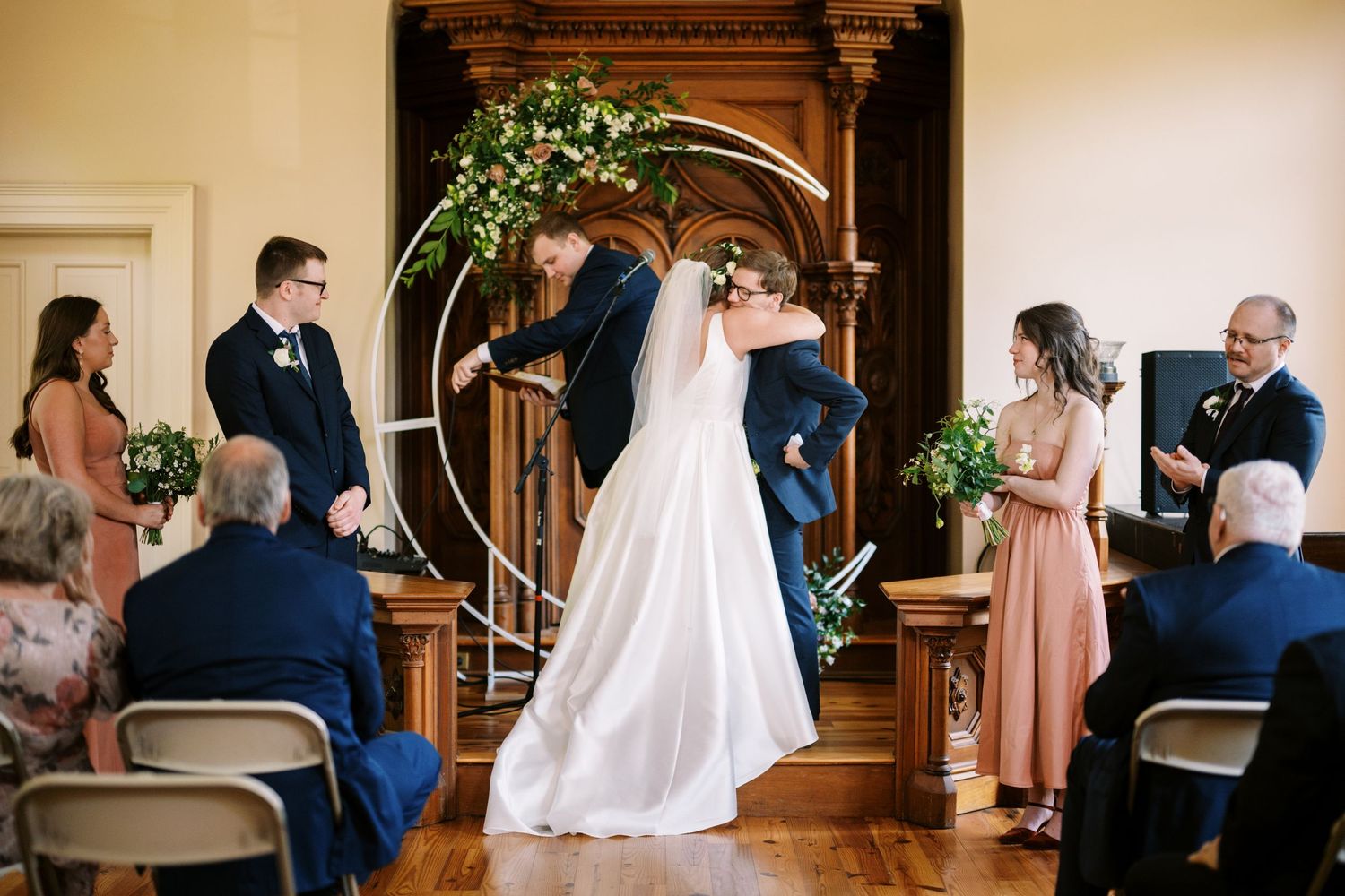 Wedding ceremony takes place in an elegant indoor venue with decorative floral arch and seated guests.