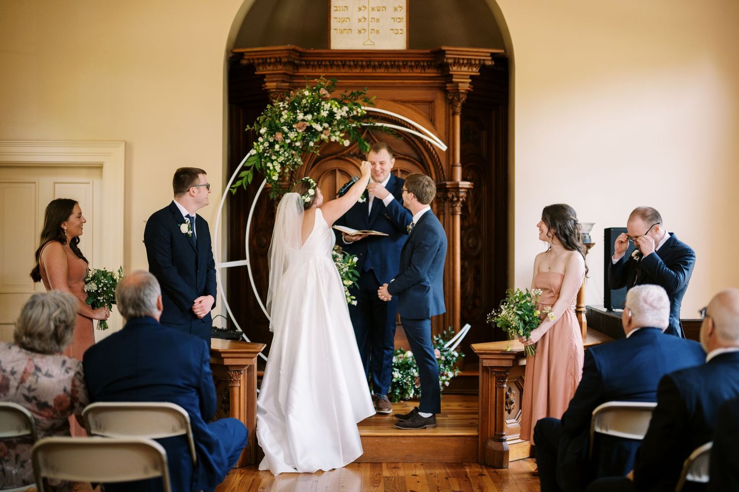 Indoor wedding ceremony with wooden arch decorated with greenery and guests seated in folding chairs.
