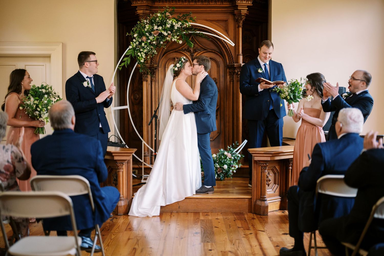 Indoor wedding ceremony in progress under wooden arch decorated with greenery at venue with hardwood floors.