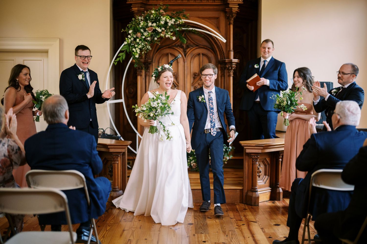 Wedding celebration under ornate wooden archway with guests watching from folding chairs.