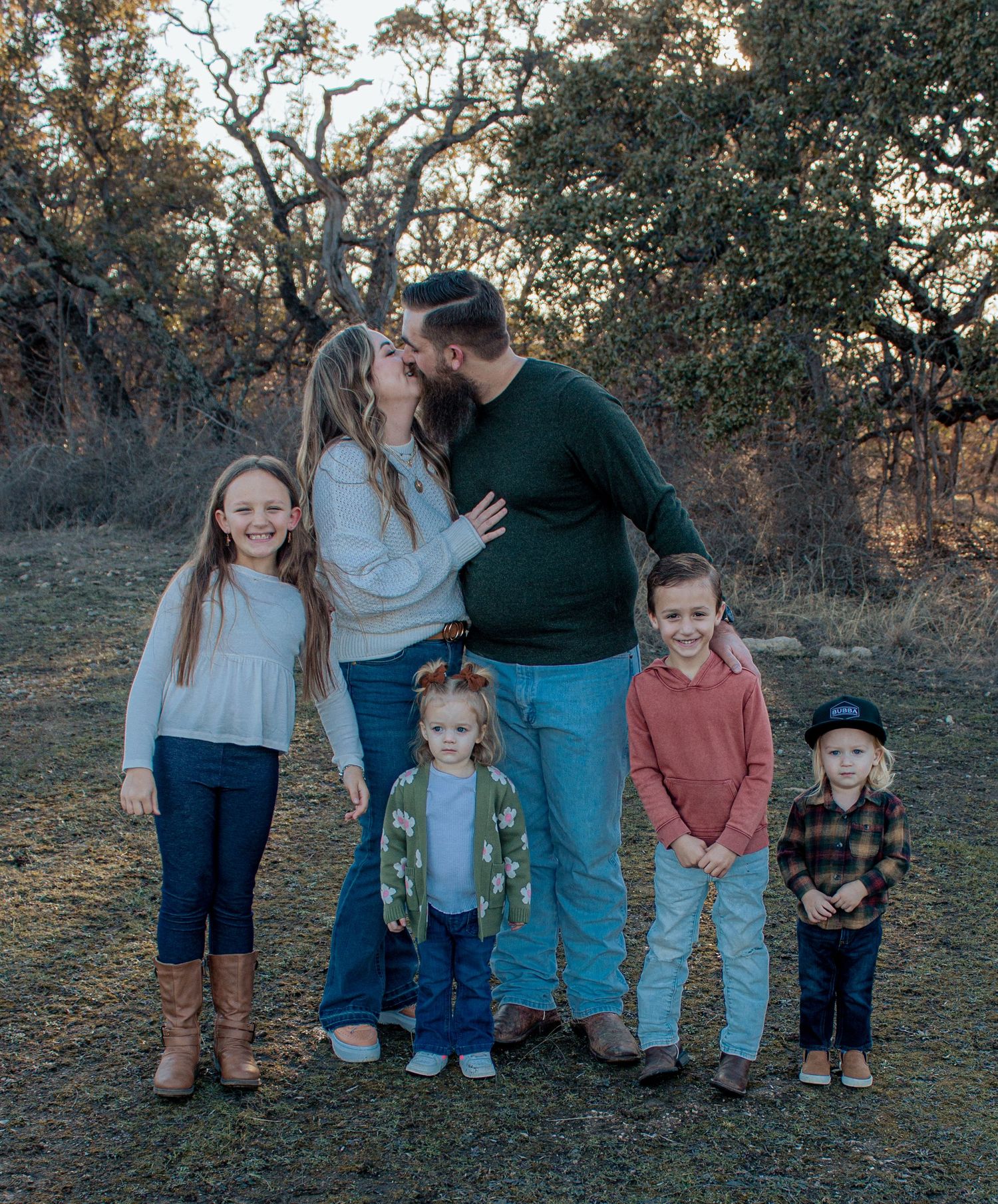 A family of five stands together outdoors at sunset among oak trees in a natural setting.