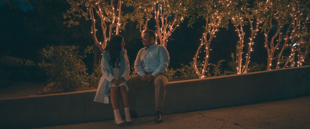 Couple sit together on a ledge at night with illuminated trees glowing warmly in the background, La La Land inspired engagement photoshoot