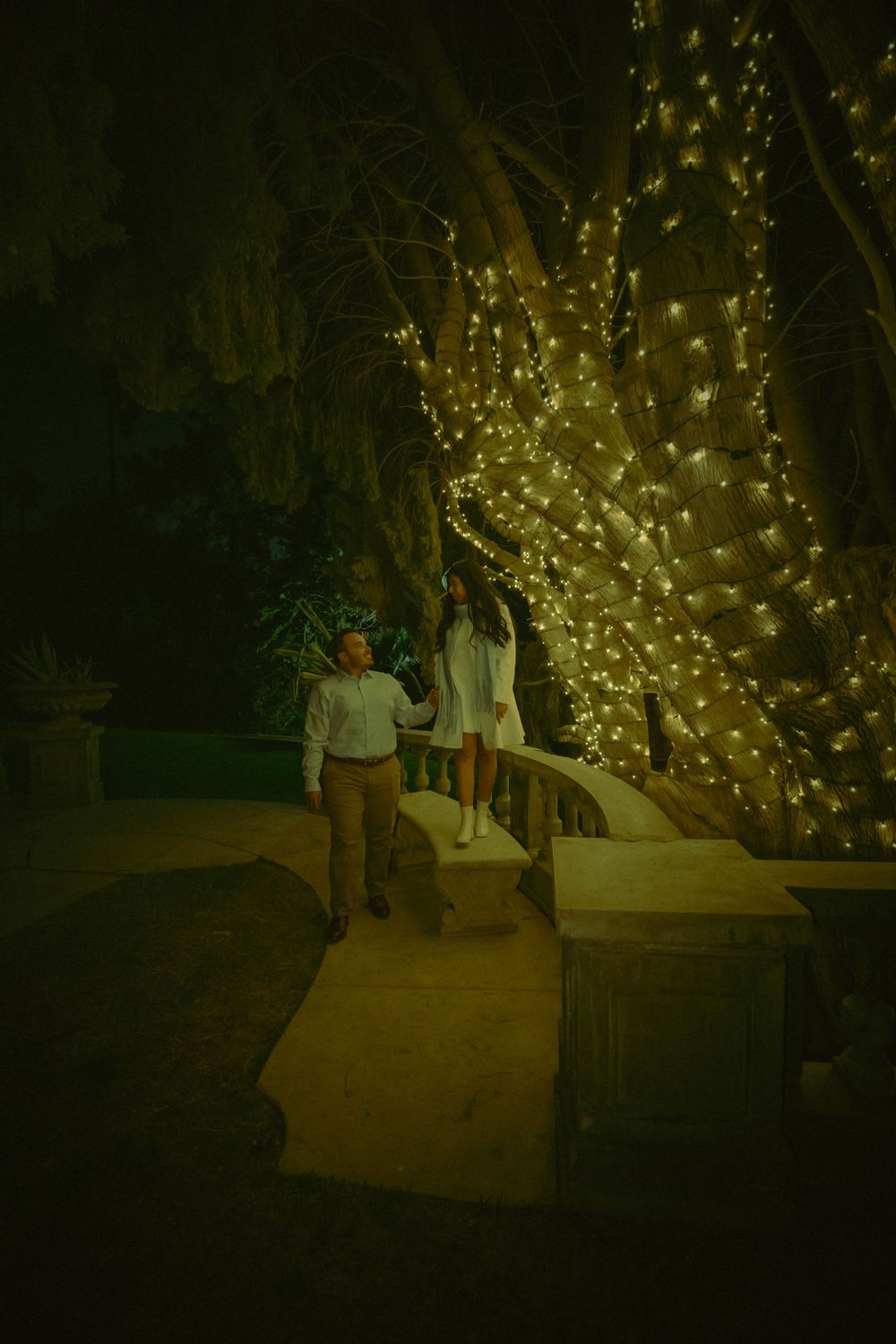 Couple walks along a curved pathway at night with illuminated trees decorated in glowing string lights creating a magical ambiance.