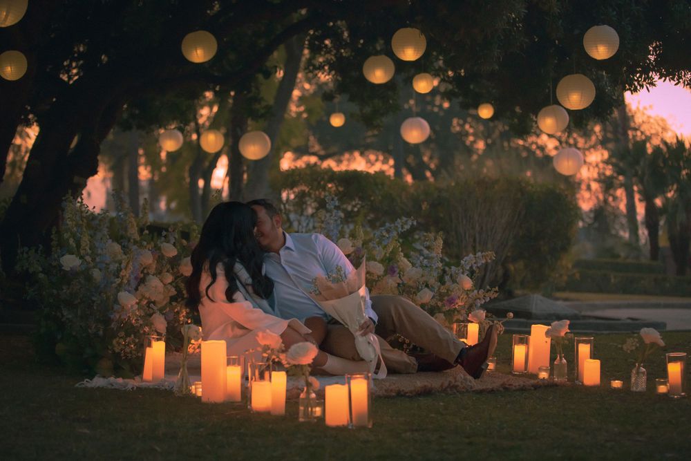 Couple sitting on blanket surrounded by glowing lanterns and paper lanterns at dusk in romantic garden setting.