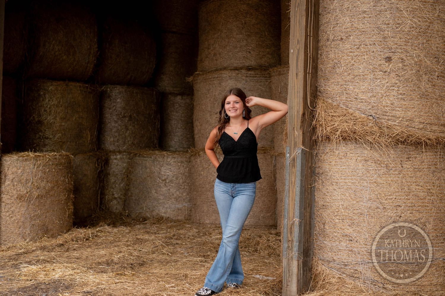 Relaxed pose against barn hay bales wearing black top and flared jeans.