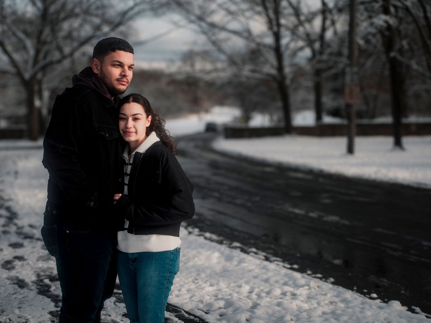 Two people embrace on a wintry road with snow-covered trees and ground in the background on an overcast day.