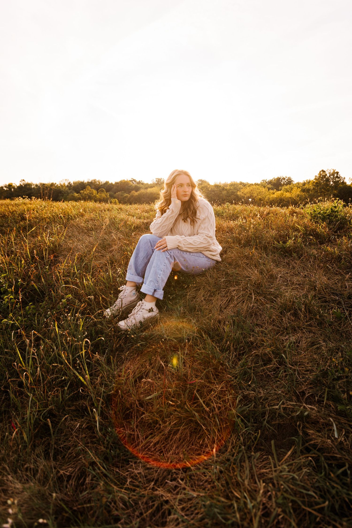 A person sitting in a sunlit golden field at sunset wearing a white sweater and jeans with boots.