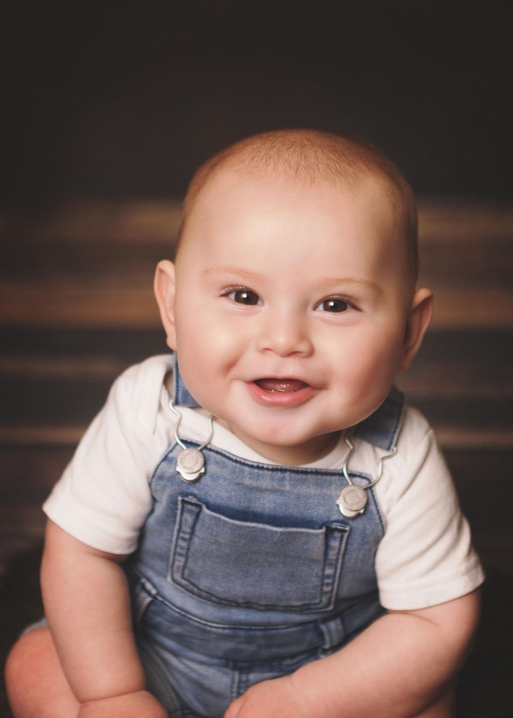Baby in denim overalls and white t-shirt smiling at camera during portrait session.