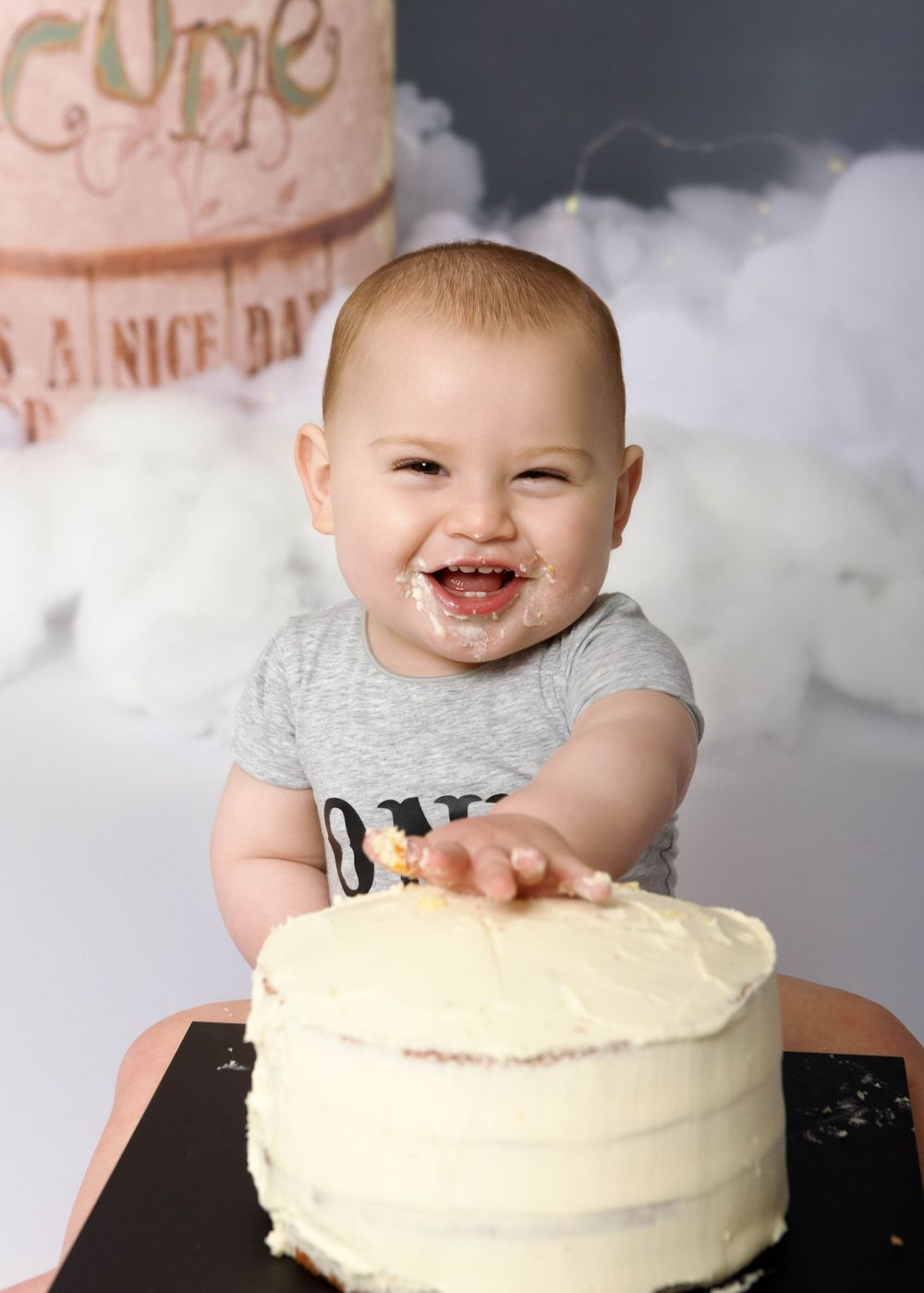 Baby in grey shirt smiling with cake smeared on face during cake smash photoshoot with white backdrop.