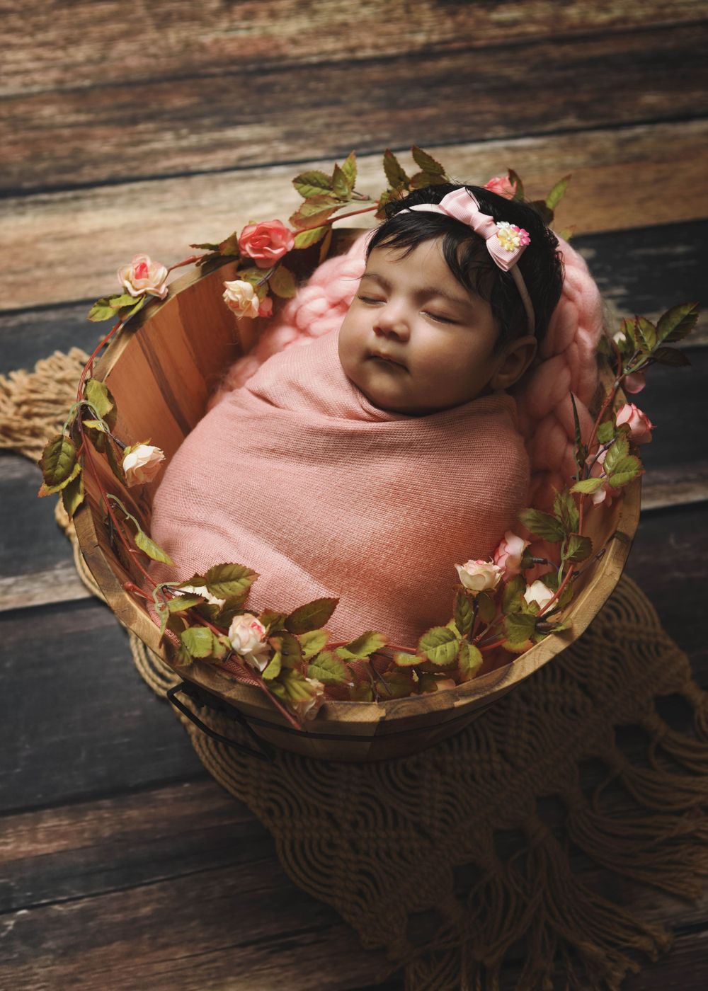 A sleeping baby wrapped in pink cloth lies in a round basket decorated with delicate pink flowers and greenery.