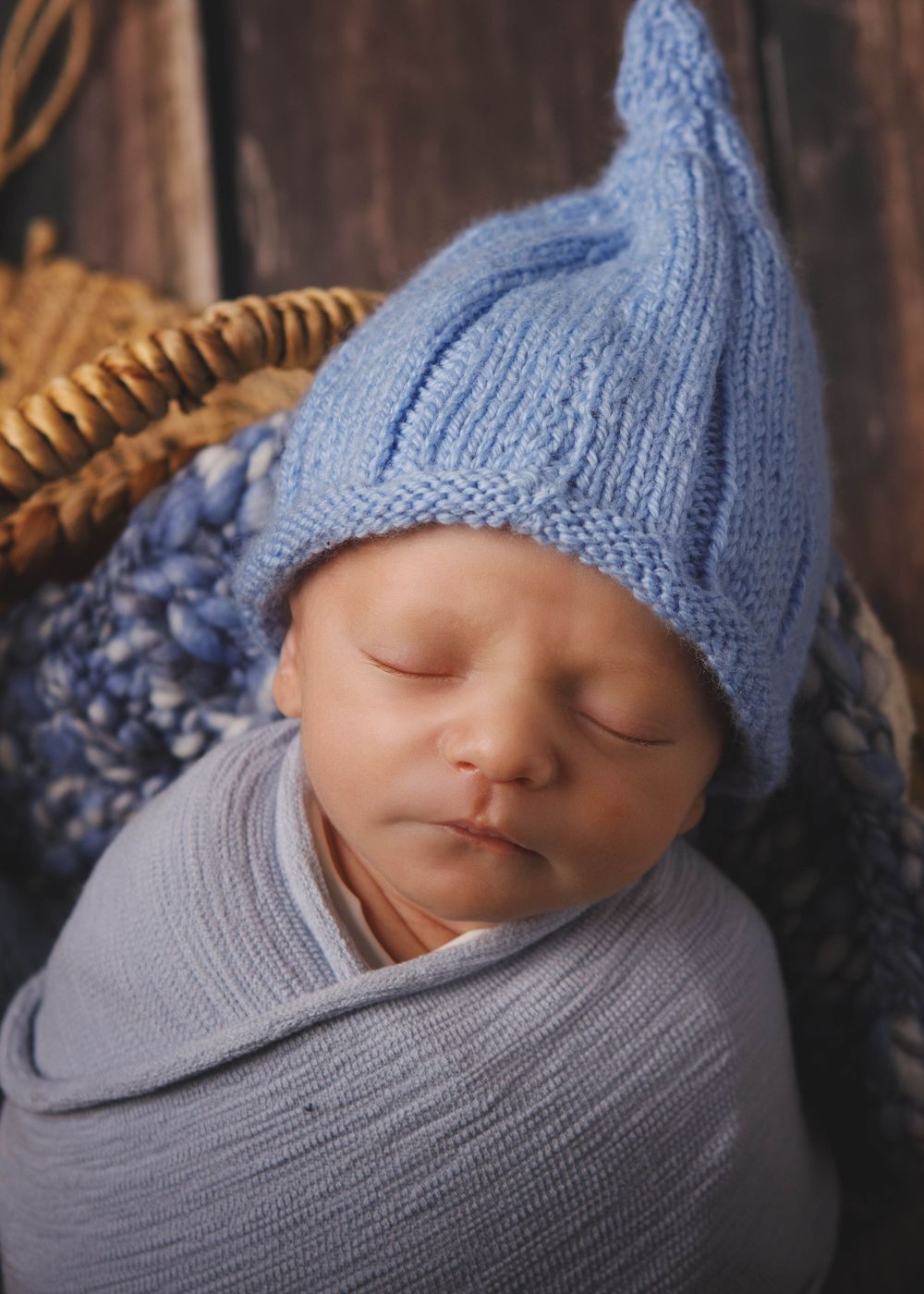 A sleeping newborn wrapped in gray fabric wearing a soft blue knitted hat with pointed top in rustic setting.