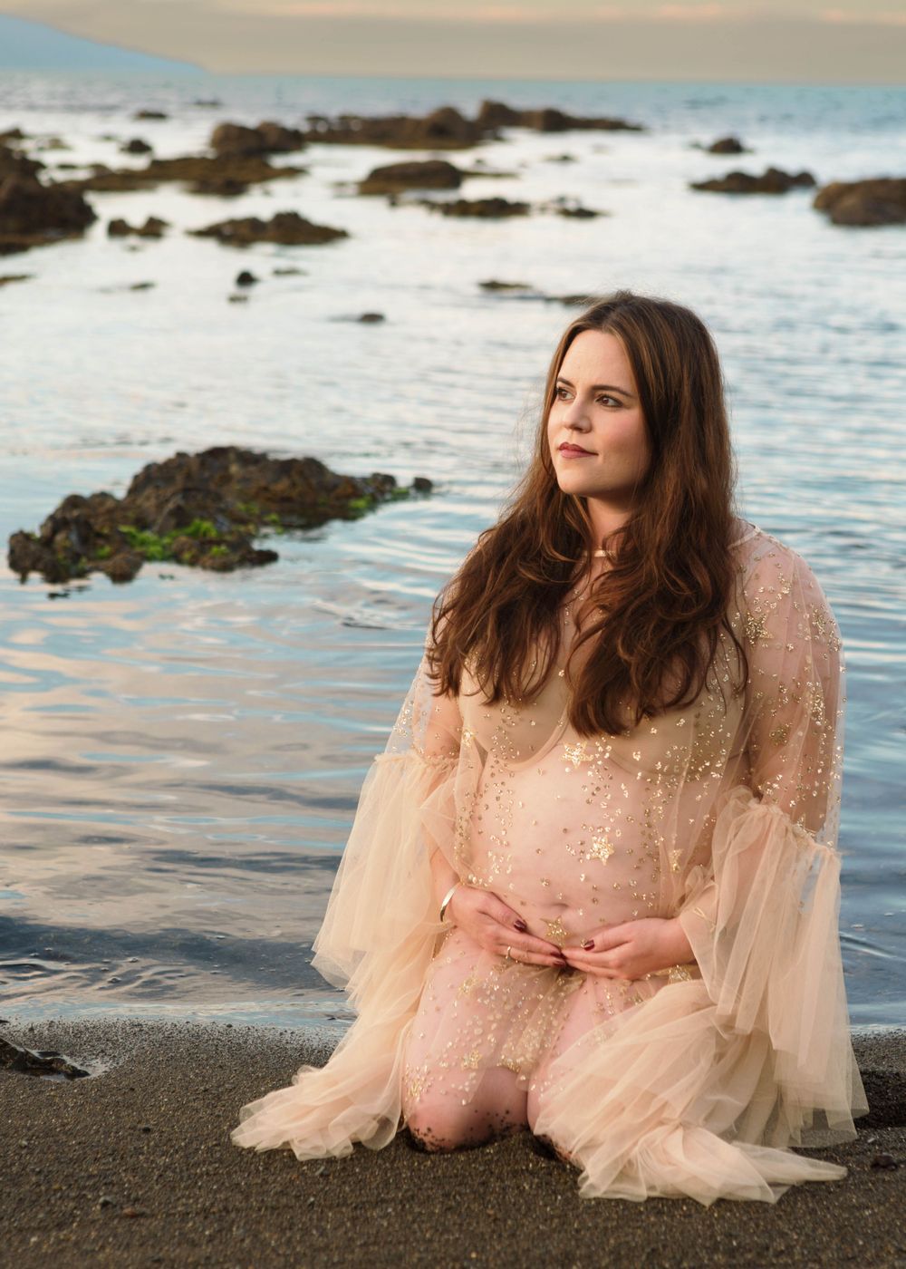 A dreamy portrait of someone in a flowing peach tulle gown sitting on a rocky beach at sunset.