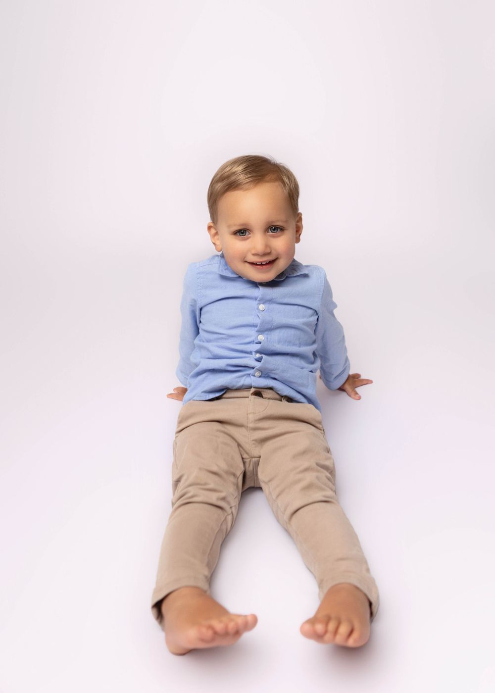 Small child wearing light blue button-down shirt and khaki pants lies on back against white studio background.