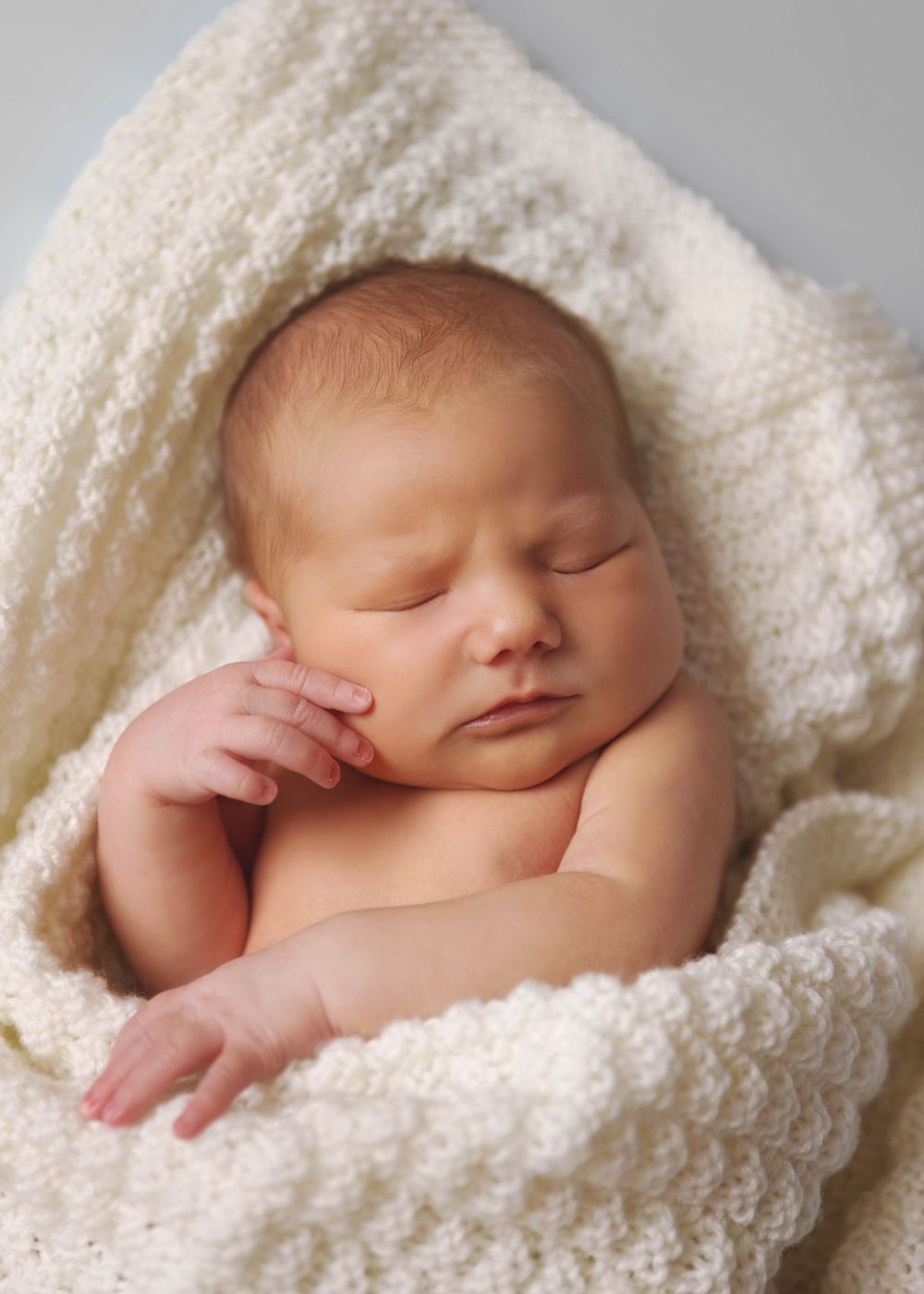 Sleeping newborn wrapped in soft white knitted blanket during professional portrait session.
