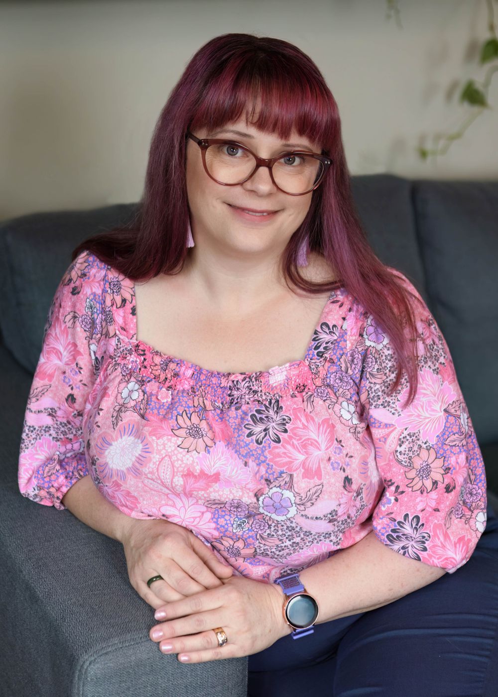 Someone with magenta hair and glasses wearing a pink floral top sits relaxed on a gray couch.