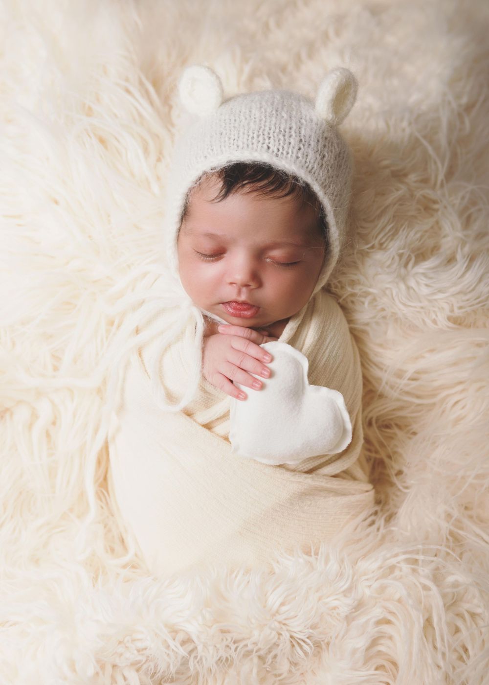 A peaceful sleeping newborn swaddled in cream fabric lies on soft white fur wearing a knitted bonnet with tiny ears.