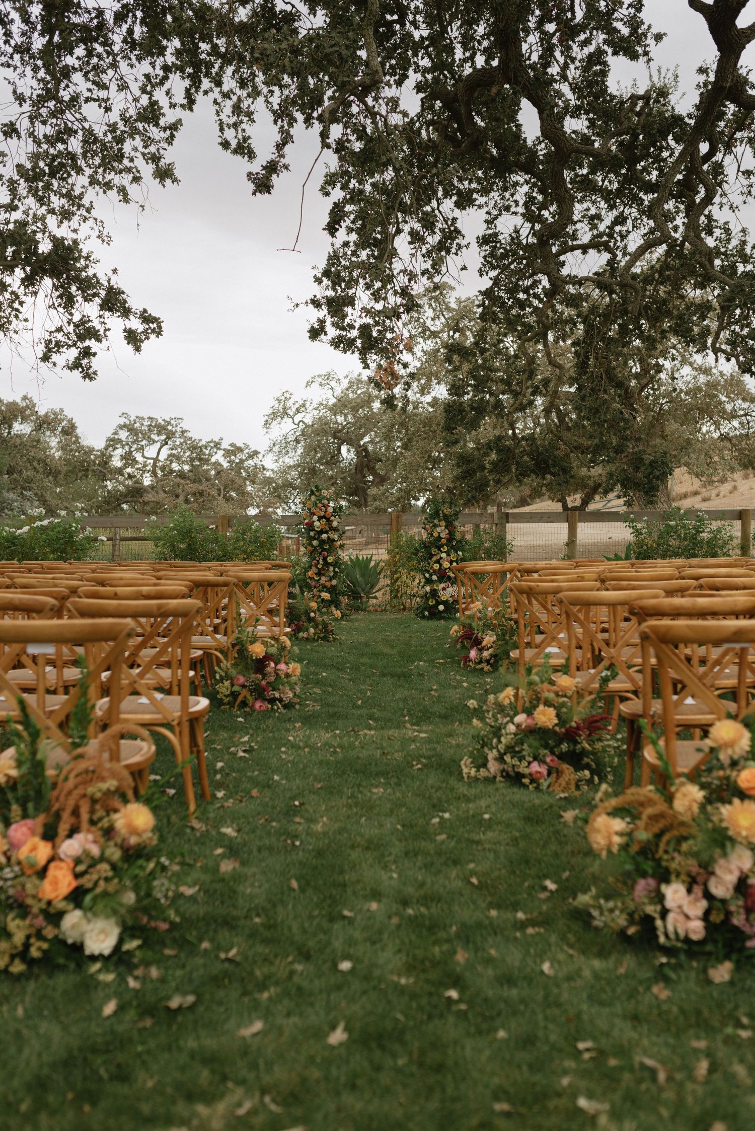 Wooden chairs line a grassy outdoor aisle decorated with peach and pink floral arrangements for a garden wedding ceremony.
