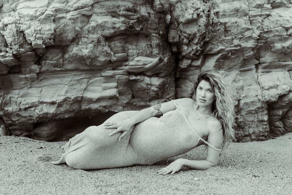 Black and white artistic maternity photo of an expecting person laying on beach sand against a rocky cliff backdrop.