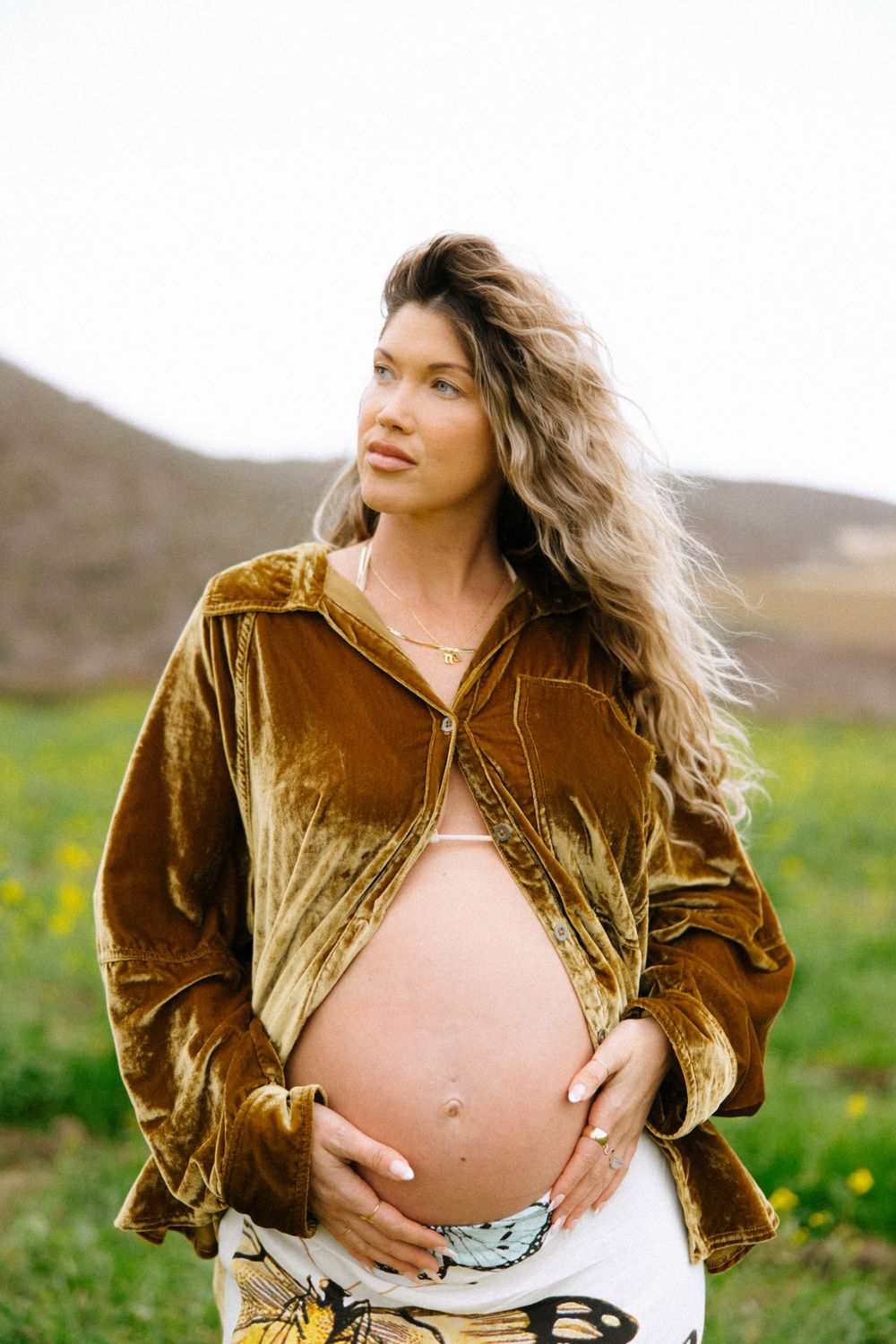 A person in a velvet gold jacket poses in a field of wildflowers, showing their pregnant belly.