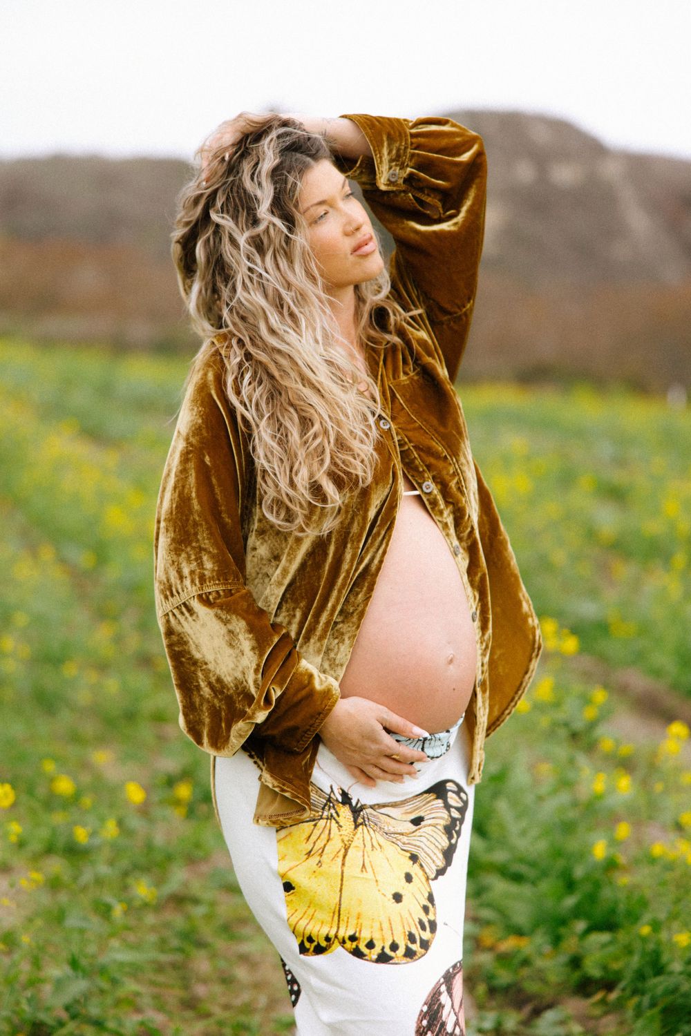 Expectant person in brown velvet coat poses in field of wildflowers with mountains in background.