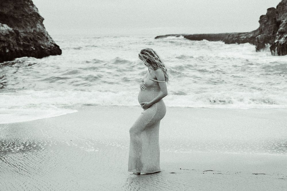 A pregnant figure in a flowing white dress stands on a beach with crashing waves and rocky cliffs in the background.