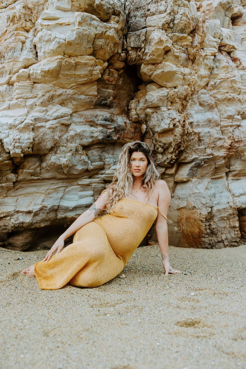 A person in a yellow dress poses on a sandy beach against a dramatic rocky cliff face in warm golden tones.